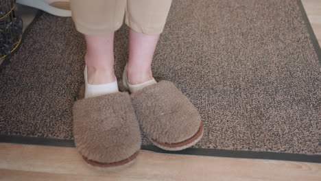lower view of guest selecting brown crocs from basket filled with slippers, wearing white socks and beige pants, preparing to put them on in indoor room with soft carpet, green wall, and wooden decor