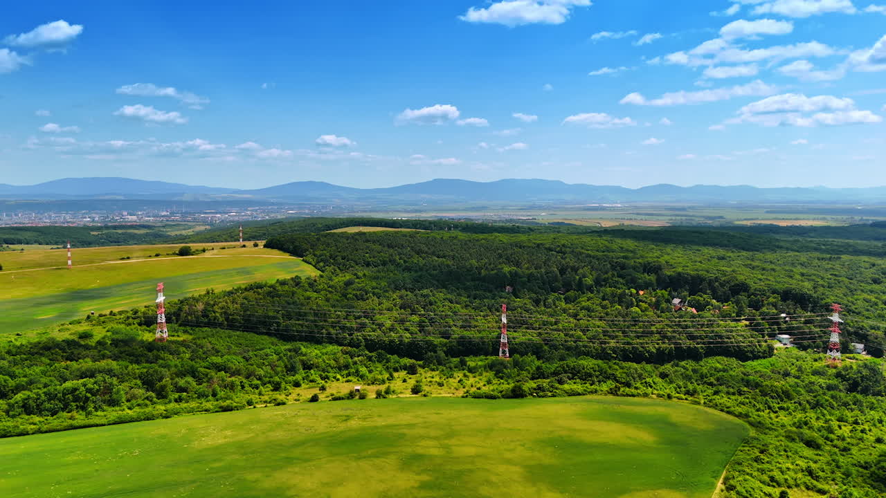 Electrical transmission towers set near the lush green forest. Green nature landscape from drone footage. Mountains silhouettes at backdrop.
