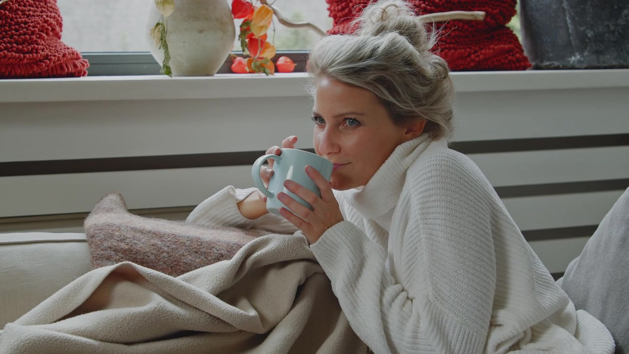 Giovane donna caucasica che si mette comoda, spensierata e sorridente, con tè caldo o caffè su un divano con una coperta accogliente durante il freddo clima invernale.