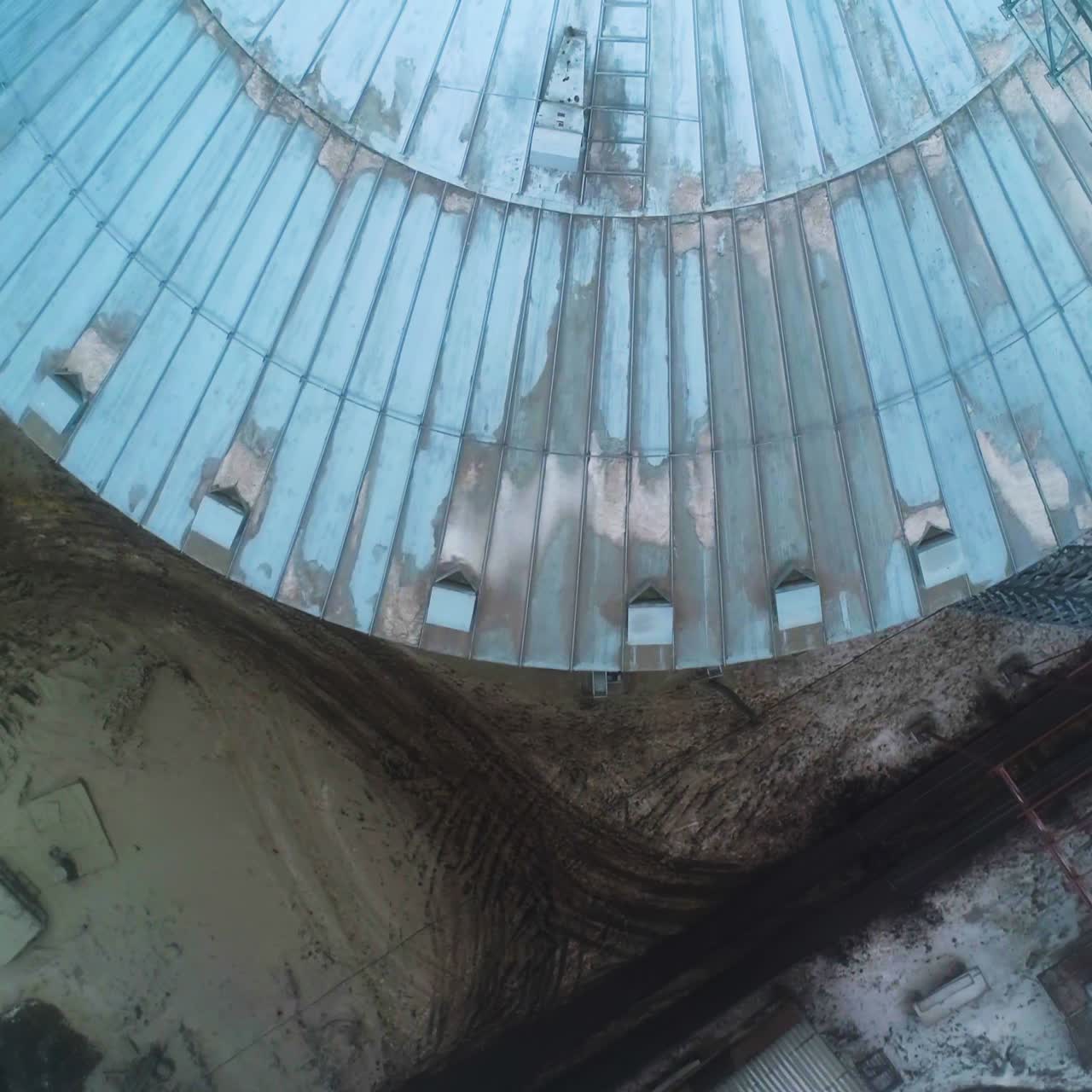 Round metal roof of silo tank. Flying over the grain container. Dirty land covered with dirty snow and trails of car wheels. Top view