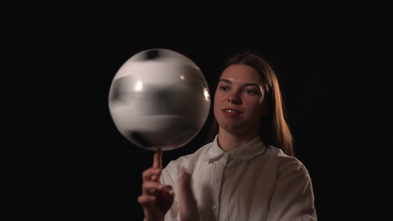 Jovem mulher de camisa branca equilibra uma tigela de vidro transparente em um dedo levantado contra um fundo escuro, criando um retrato focado e elegante.