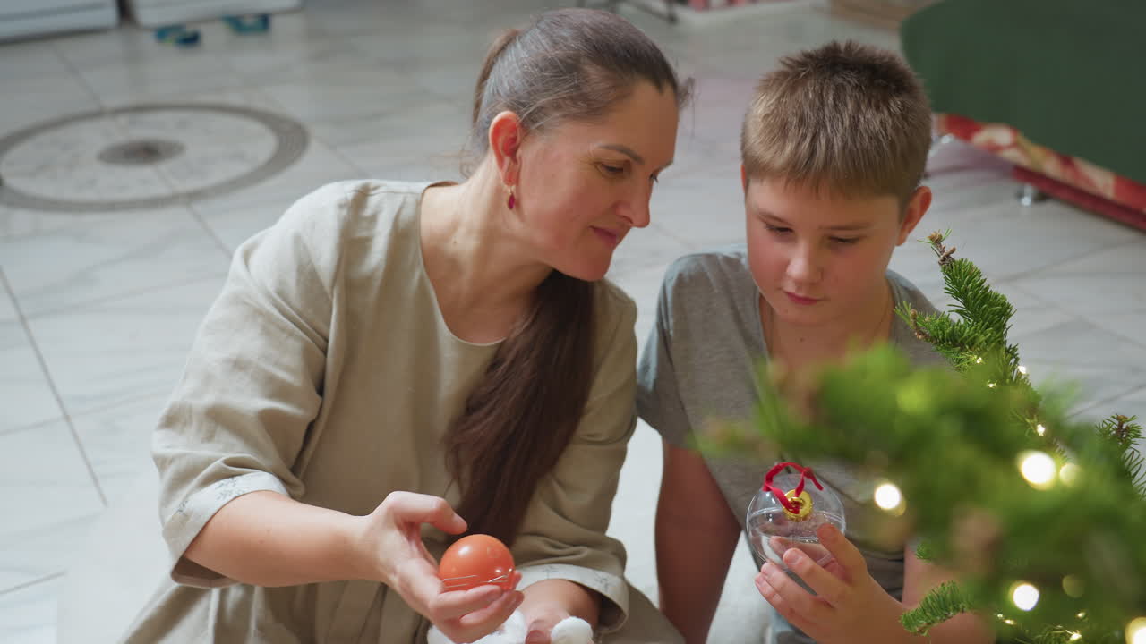 Mom and son sitting together indoors looking at Christmas ornament in their hands, cozy festive atmosphere with Christmas tree decorated with glowing lights in background, capturing joyful holiday