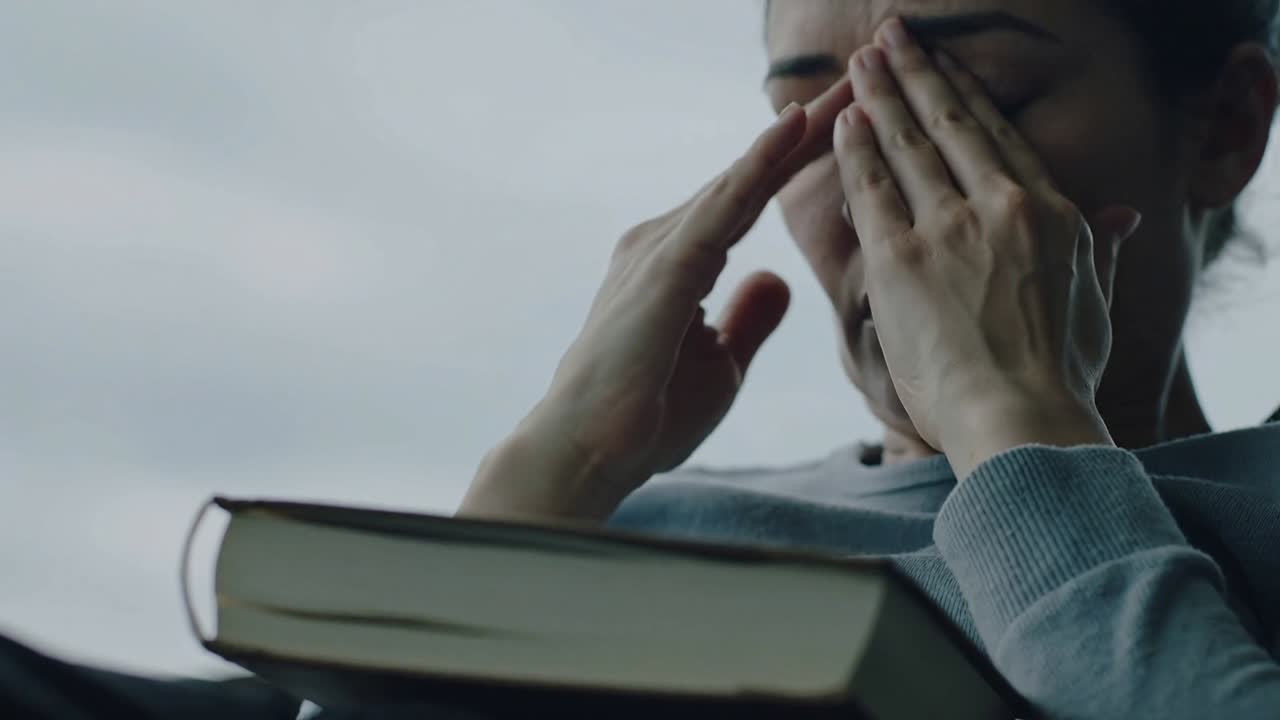 Donna con maglione azzurro che si strofina dopo aver letto un libro con copertina rigida sul divano vicino alla finestra, con un segnalibro a nastro.