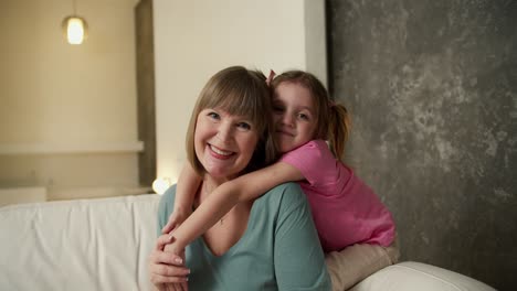 Mature-loving-grandmother,-her-adorable-little-granddaughter-hugs-her---photoshoot-for-camera-seated-on-sofa-in-living-room