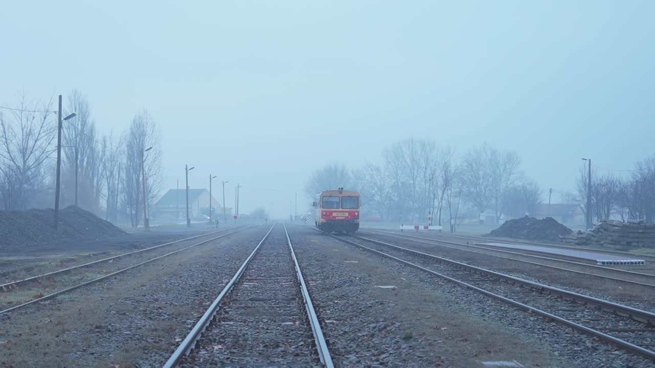 A Hungarian passenger train leaves a small rural station on a misty day, traveling along empty railway tracks