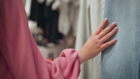 hand view of a lady in a pink shirt walking through a clothing store as she touches blue and ash jeans hanging on the rack, feeling their texture, blurred view of other clothes in the background