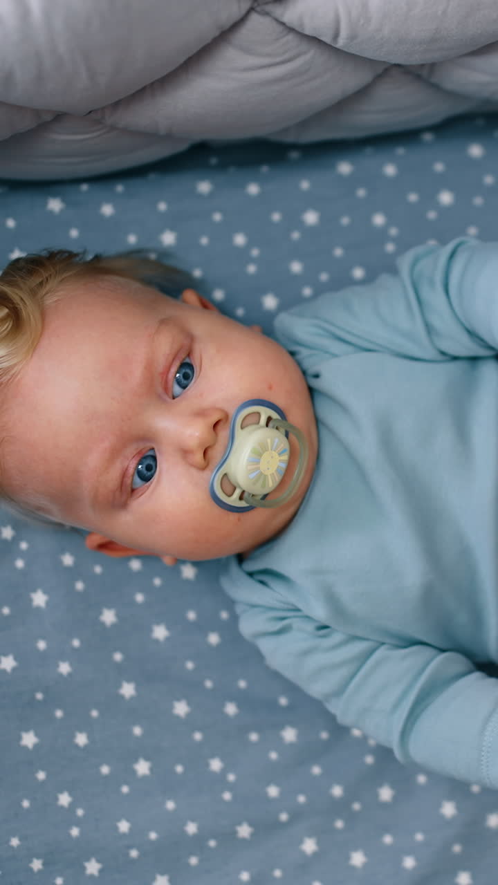 Lovely plump baby boy with pacifier lies in the crib holding legs up. Kid looks on the cot mobile hanging above. Top view. Vertical video