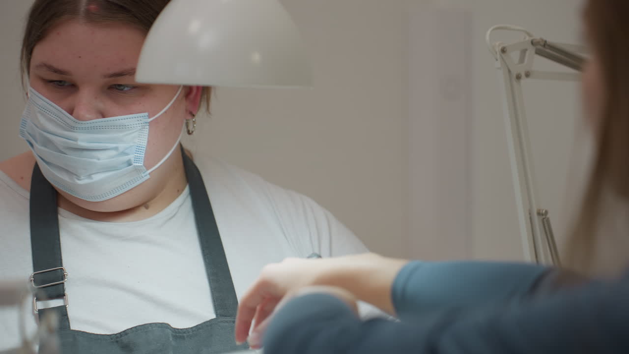 Focused nail technician in mask and black gloves using electric filer on customer nails while holding small scissors and cleaning brush under bright lamp during detailed manicure procedure