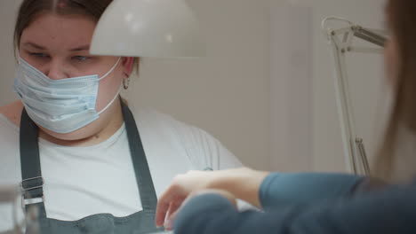 focused nail technician in mask and black gloves using electric filer on customer nails while holding small scissors and cleaning brush under bright lamp during detailed manicure procedure