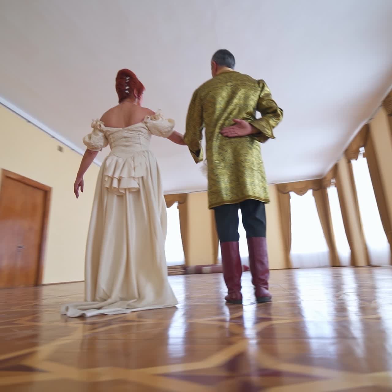 Woman in a long white historic dress dances to a man wearing old golden jacket. Circling footage around the aging couple performing old type dance. Low angle view