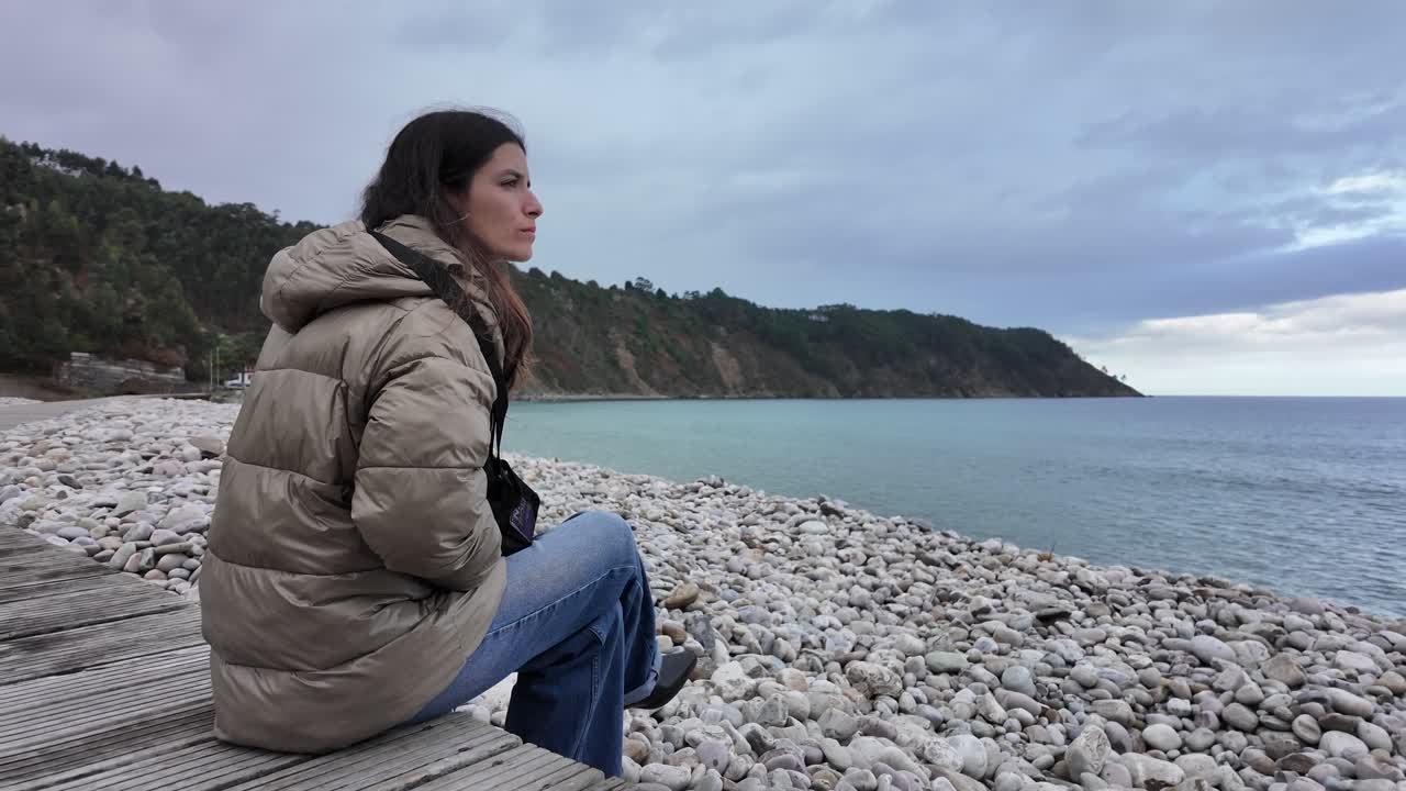 Slow-motion shot of a woman sitting quietly by the sea on a cold day