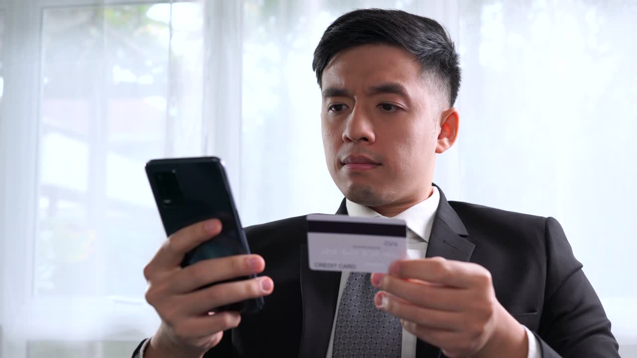 Pengusaha muda Asia yang cerdas duduk di kantor rumah dengan laptop, tersenyum dan bahagia saat menggunakan kartu kredit dengan aplikasi belanja online di ponsel pintar