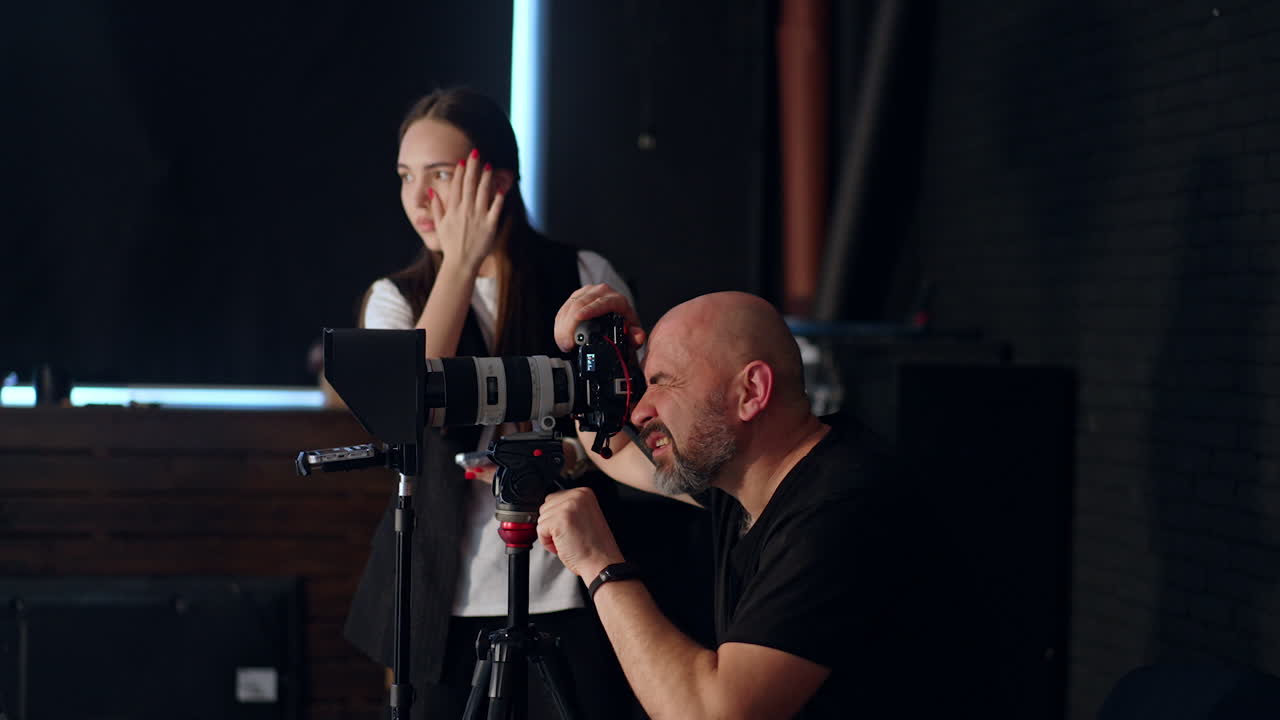 Bald bearded photographer looks at camera on tripod. Girl at backdrop uses a smartphone. Photo studio footage backstage.