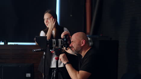 bald bearded photographer looks at camera on tripod. girl at backdrop uses a smartphone. photo studio footage backstage.