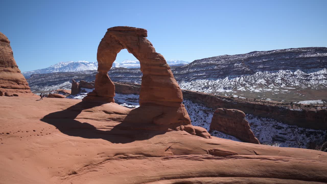 Pendaki berjalan mendekati dan memanjat Delicate Arch di Taman Nasional Arches, gerakan panning.