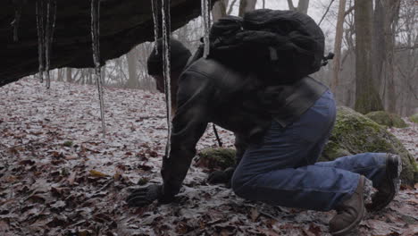 a-bearded-hiker-wearing-a-plaid-flannel-shirt-and-backpack-climbs-to-the-ground-to-enter-a-cave-under-the-rocky-ledge-with-icicles-hanging-from-the-rocks-in-winter-in-Ohio