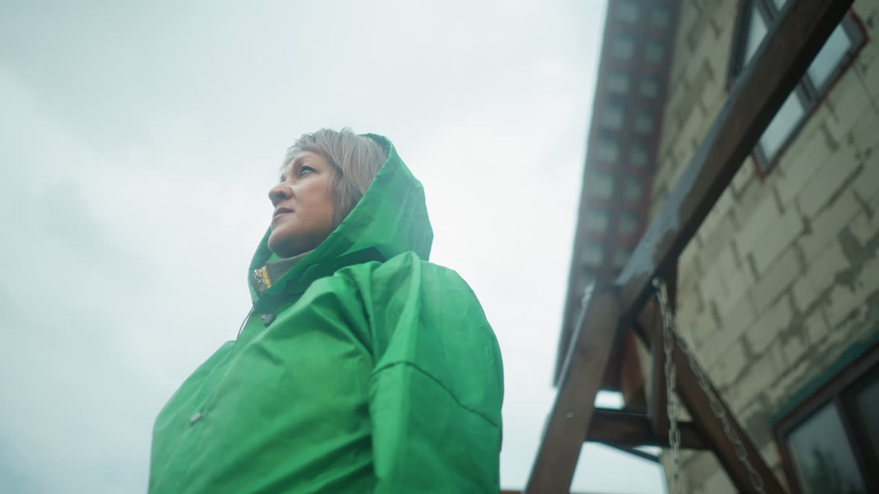 밝은 하늘 아래 초록색 레인코트를 입은 여성의 클로즈업, 그녀가 천천히 오른쪽으로 돌면서 초점을 맞추고, 스 벤치와 건물의 부분적인 시각이 배경에 나타납니다.