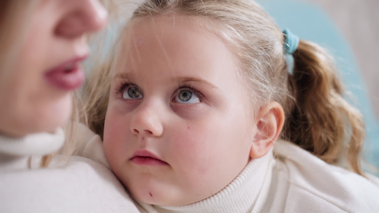 Close up of young girl with blonde hair tied in ponytail wearing white turtleneck looking up at woman with wide eyes full of curiosity and innocence, tender family moment with soft emotional focus