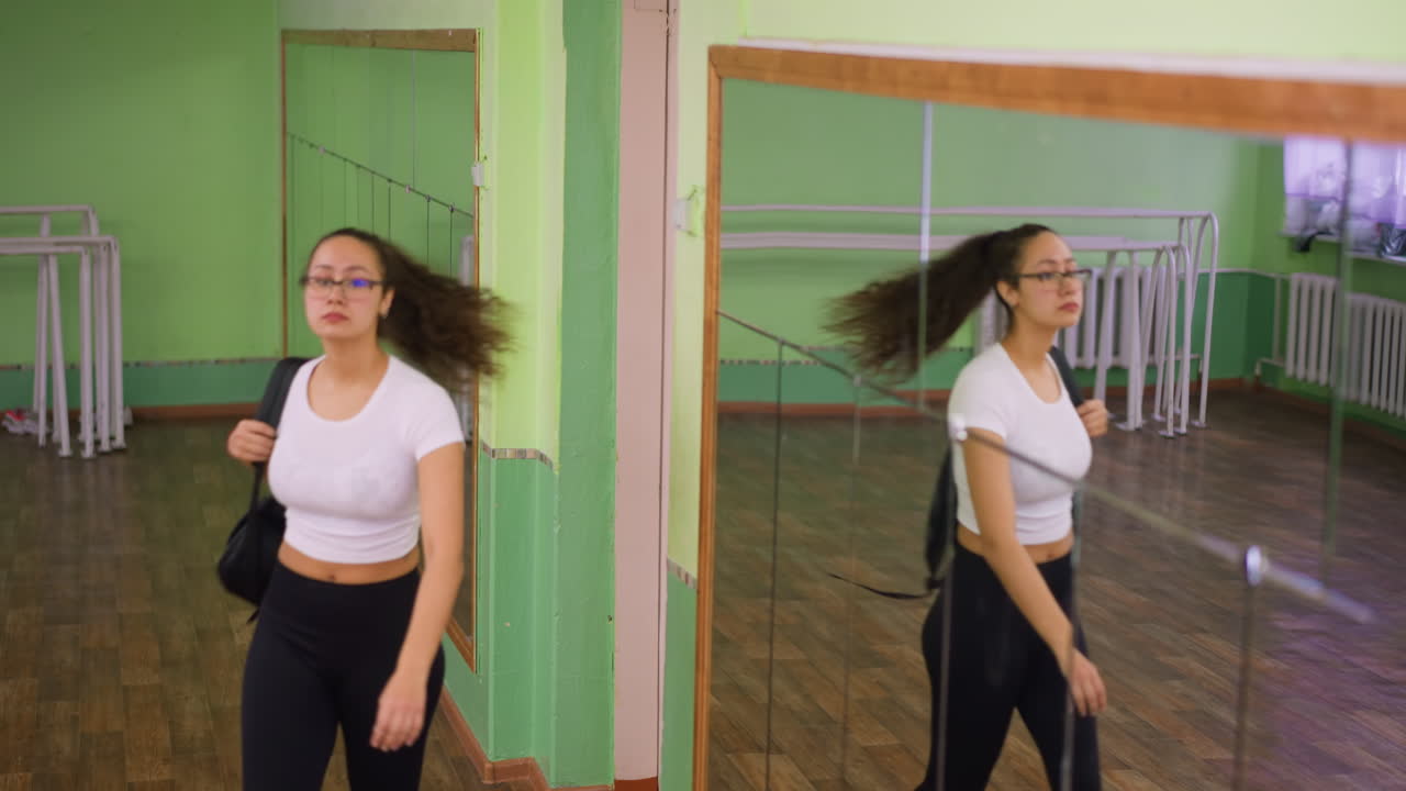 Back view of woman in white top and black leggings carrying school bag closing door calmly then turning back to walk with background mirror reflecting her body in bright indoor hall