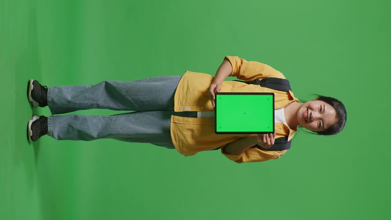 Full Body Of Asian Woman Student With A Backpack Smiling And Showing Green Screen Tablet To Camera While Standing In The Green Screen Background Studio