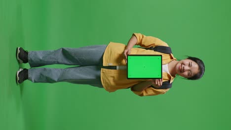 full body of asian woman student with a backpack smiling and showing green screen tablet to camera while standing in the green screen background studio