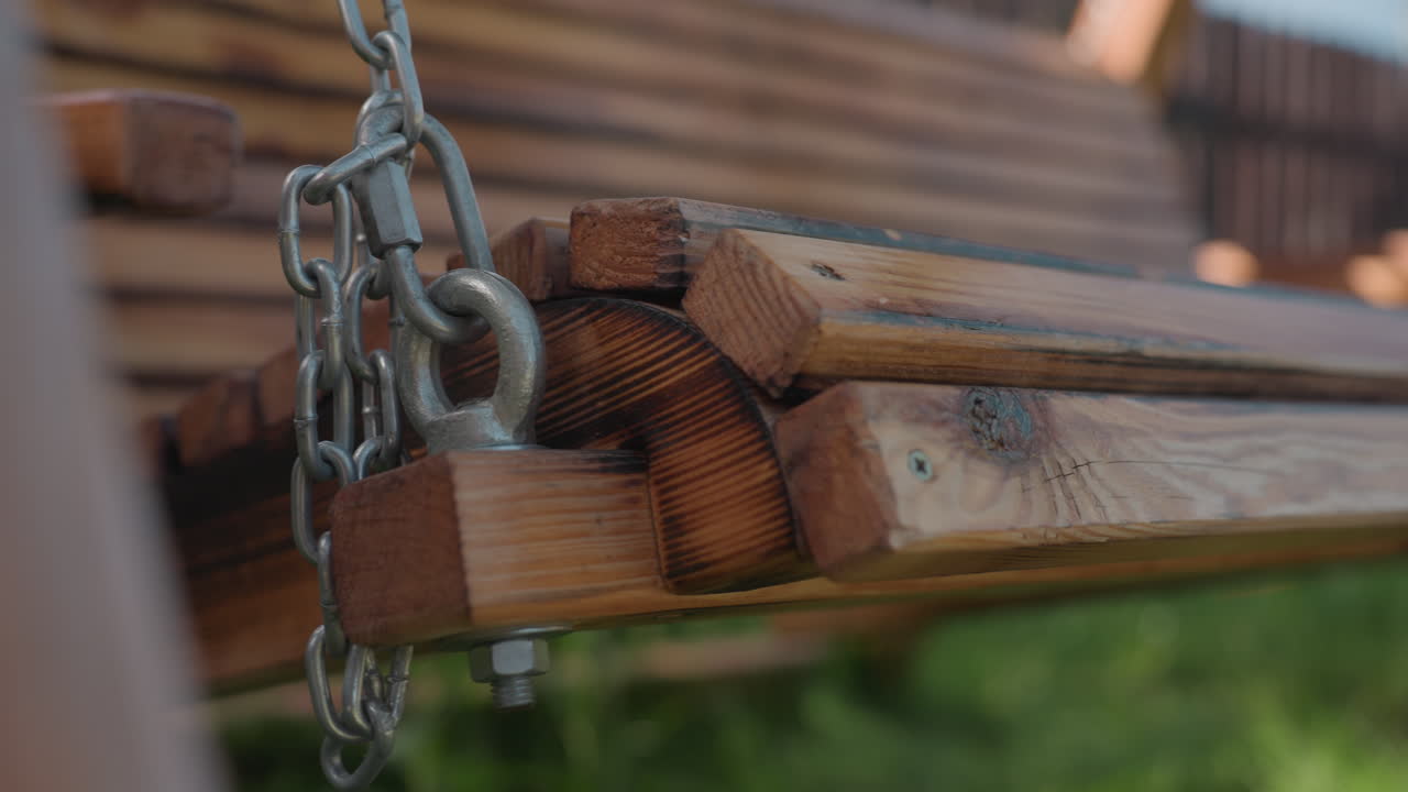 Close up of strong metal chain connected to sturdy wooden joint of outdoor swing, highlighting craftsmanship and smooth motion of bench seat swaying gently in sunlight over green grass in backyard