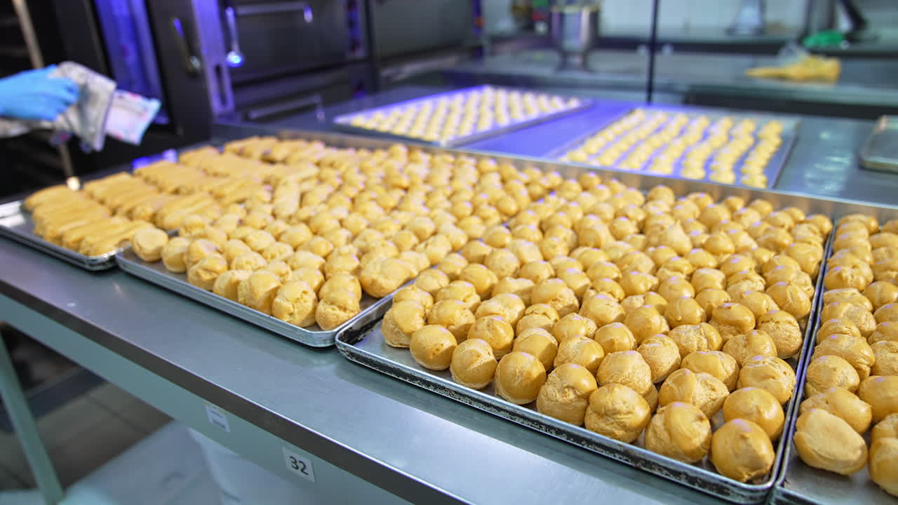 Large metal tables covered with baking sheets full of baked éclairs. Pastry prepared for stuffing with cream. Food factory production.