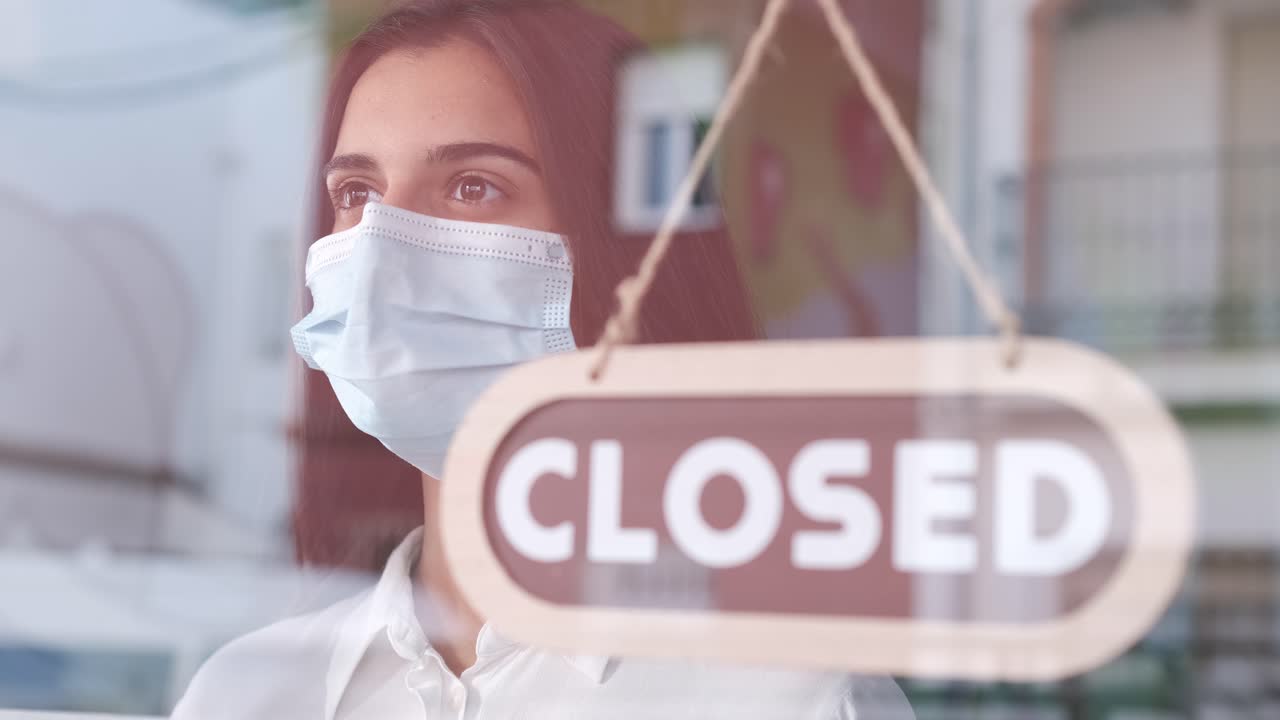 Girl with face mask changing closed to open sign on window looking outside waiting for clients. Start-up at covid time concept.