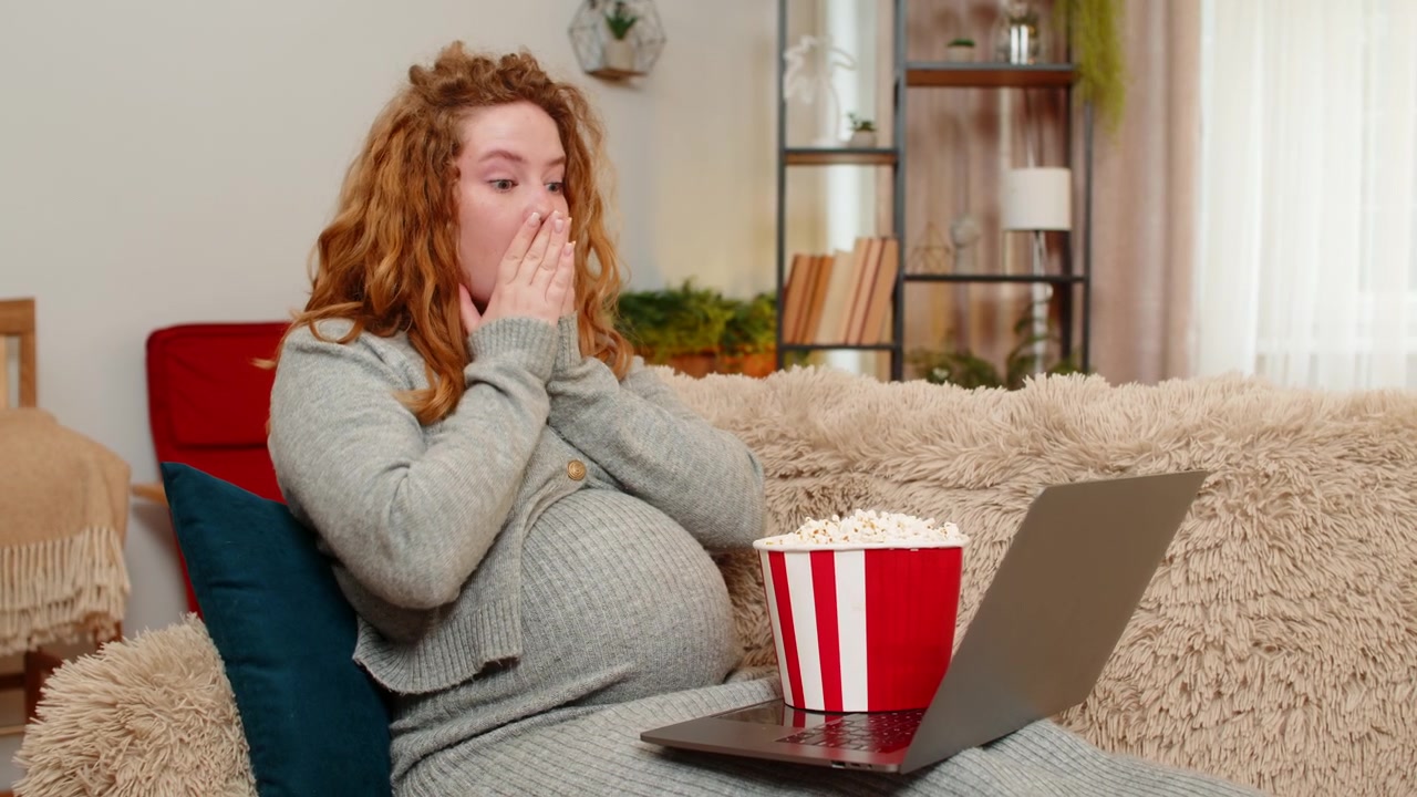 Donna incinta che guarda un film horror sul laptop a casa, coprendosi gli occhi, mangiando popcorn, ansiosa e divertita.