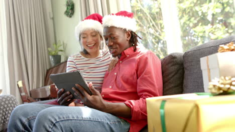 Feliz-Pareja-Diversa-Con-Sombreros-Navideños-Usando-Tableta-En-Una-Sala-De-Estar-Soleada,-Cámara-Lenta