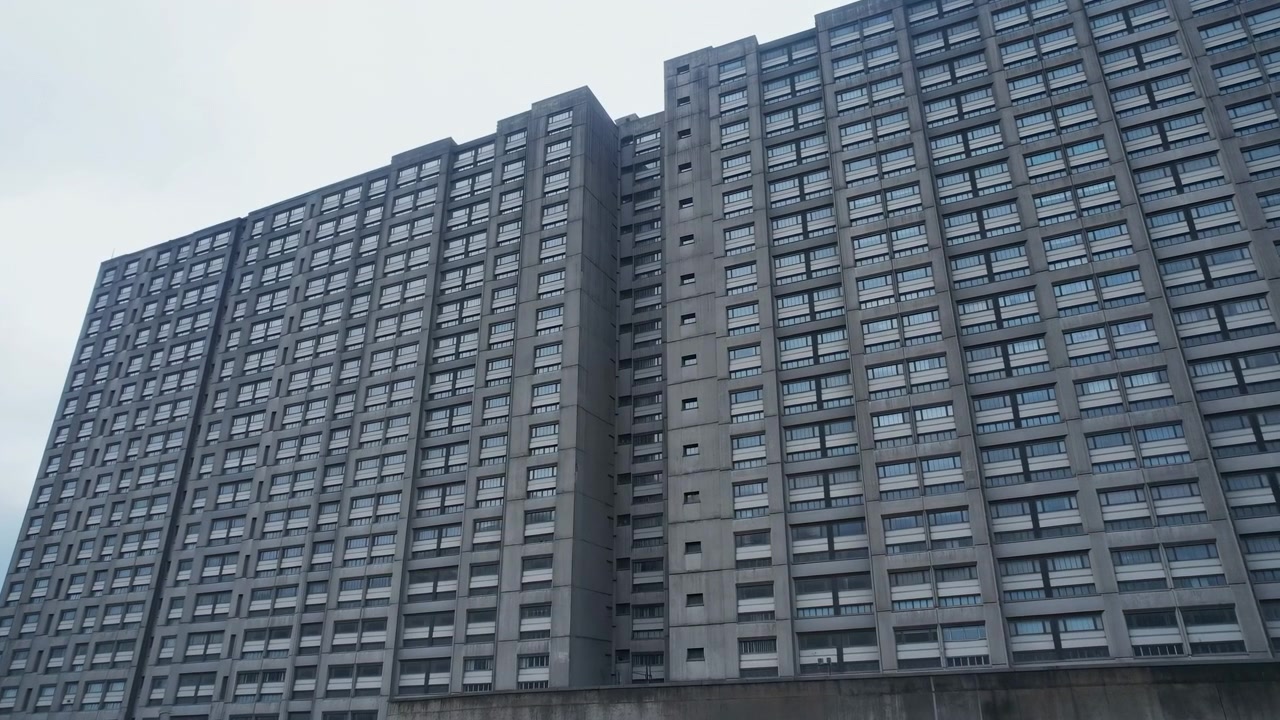 O plano médio retrata um imponente edifício residencial cinza com inúmeras janelas e painéis de concreto, apresentando uma fachada urbana expansiva contra um céu nublado.