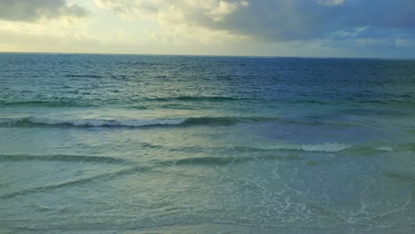 ocean waves arriving at low tide on a sandy beach under a cloudy sky