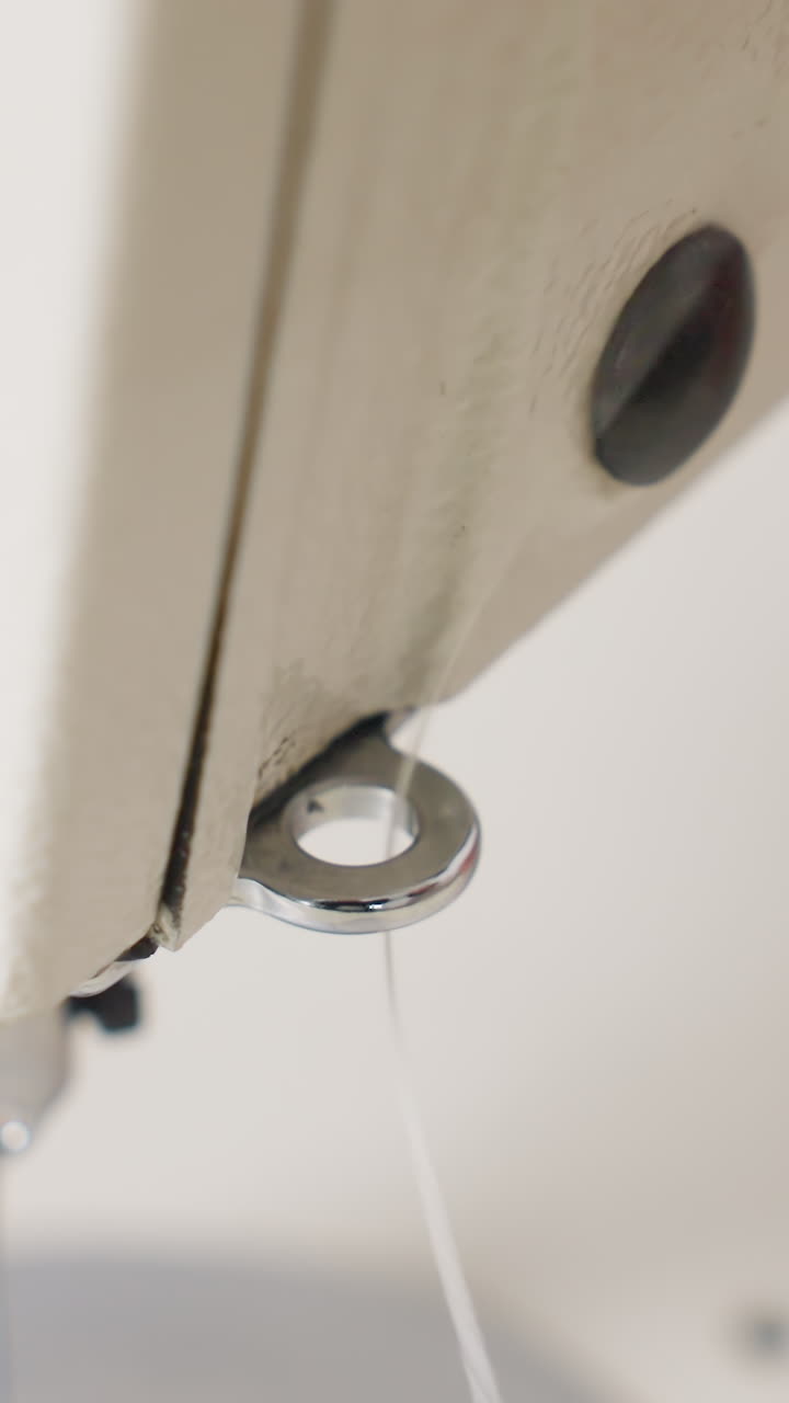 closeup hand threading machine eyelet near throat plate, hobbyist pulling white thread through circular metal guide, subtle relief as thread seats cleanly, neutral background, intimate DIY workshop