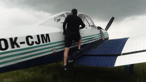 following fhd shot of a man climbing on a wing of a small sport airplane, preparing to enter the cabin and take off from a field airport with a grey stormy sky above