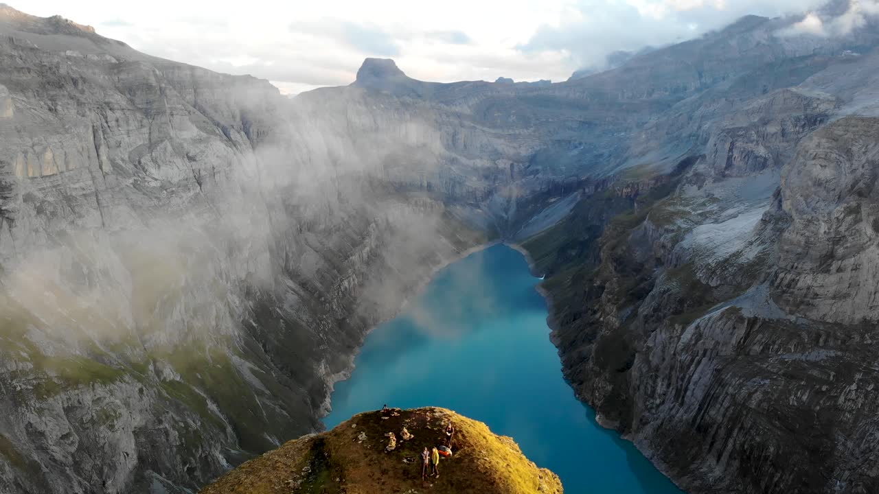 Sorvolo aereo da un punto panoramico del Limmernsee a Glarona, Svizzera, attraverso nuvole incandescenti con escursionisti che si godono la vista delle scogliere, del paesaggio e del lago delle Alpi Svizzere dal loro campeggio al tramonto