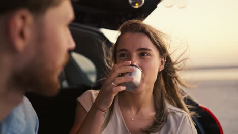 pov: blonde girl listening to her bearded boyfriend while sitting in the trunk of a black car, drinking tea and laughing against