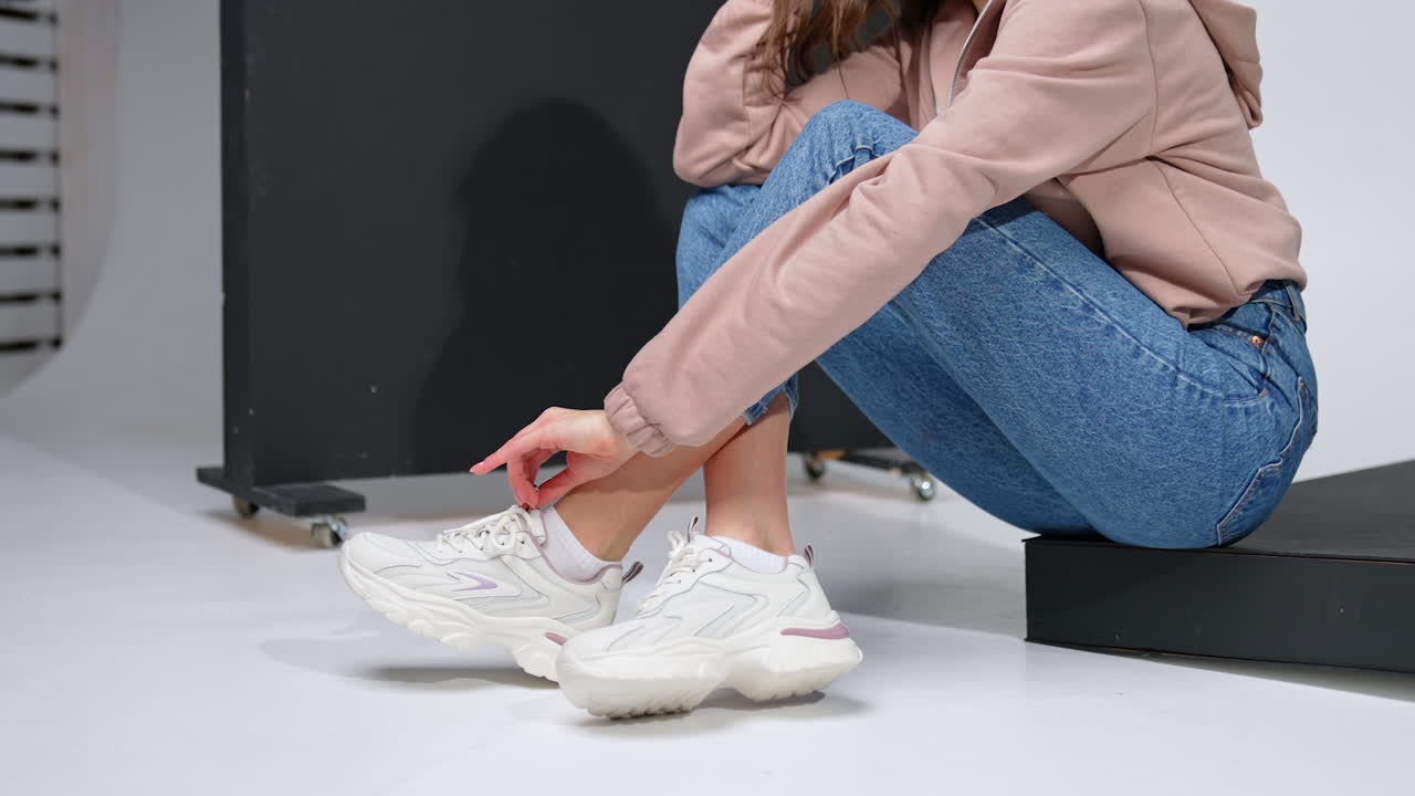 Cropped image of a girl sitting on a black step. Model in jeans, sweatshirt and white sneakers in studio.