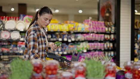 A-confident-brunette-girl-in-a-checkered-shirt-is-driving-a-cart-in-a-supermarket-and-choosing-an-avocado-in-the-fruit-department-of-the-supermarket