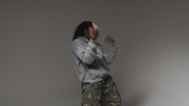 Studio Portrait Shot Of Young Woman Wearing Hoodie Dancing With Low Key Lighting Against Grey Background