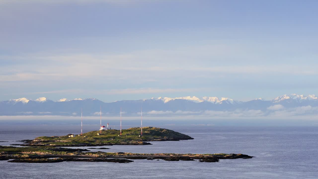 A serene coastal landscape with a small island, lighthouse, and antennas. Snow-capped mountains and a calm sea under a clear blue sky complete the scene