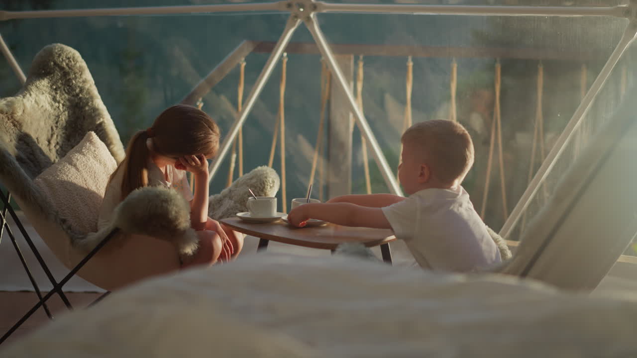 아이들은 창문 의 테이블에 앉아 있습니다. 소년은 빈 컵을 밀어 니다. 형제 자매는 핑 사이트의 쾌적한 텐트에서 평화로운 순간을 즐기고 있습니다. 아이들은 호텔 부지에 있습니다.