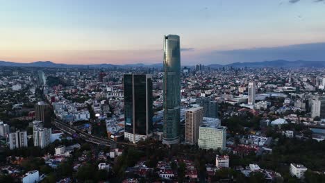 Aerial-view-circling-around-the-Torre-Mítikah-Tower,-vibrant-dusk-in-Mexico-city