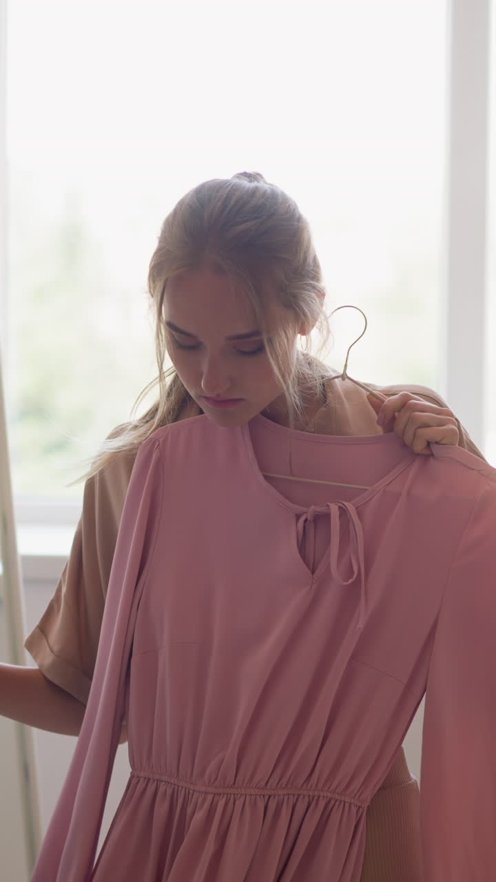Young blonde woman chooses between elegant pink and classic black dresses for perfect evening outfit in fitting room at home slow motion