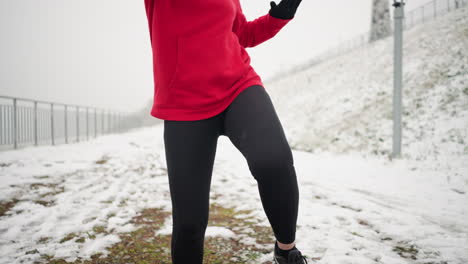 close-up of woman exercising outdoors, lifting her leg during workout on snowy ground with a snowy hill and iron railing in the background, dressed in a red hoodie and black leggings