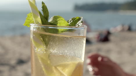 Close-up-of-hand-decorating-drink-with-fresh-mint-herb-and-being-taken,-sunny-beach-in-background,-Slowmo