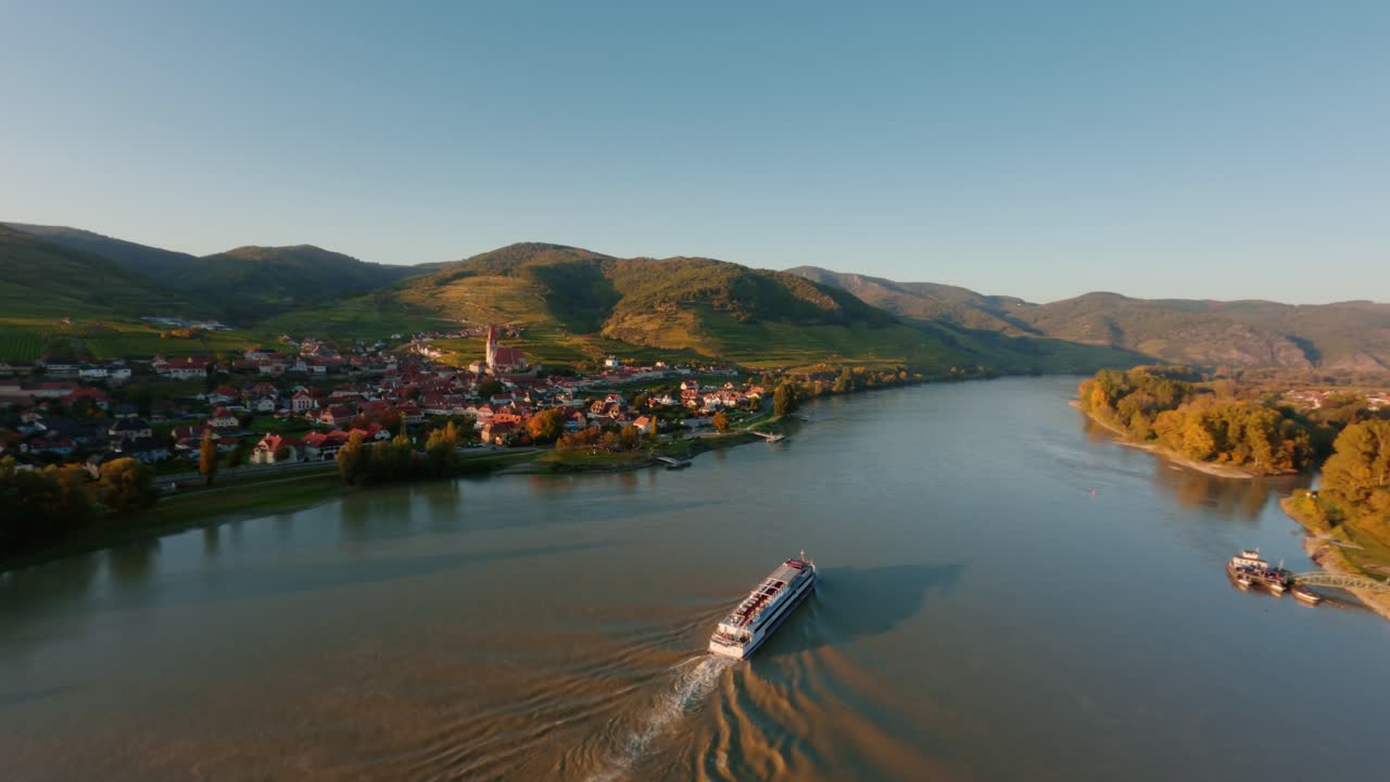 Slow ascending FPV drone shot of a danube boat passing Weissenkirchen town in wachau valley dureing sunset time in autumn