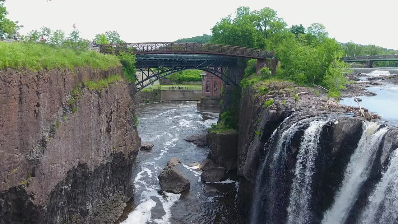 A spectacular 4K drone shot of the historic Paterson Great Falls, located in Paterson, New Jersey. The camera moves forward over the waterfall and under a walking bridge crossing the Passaic River