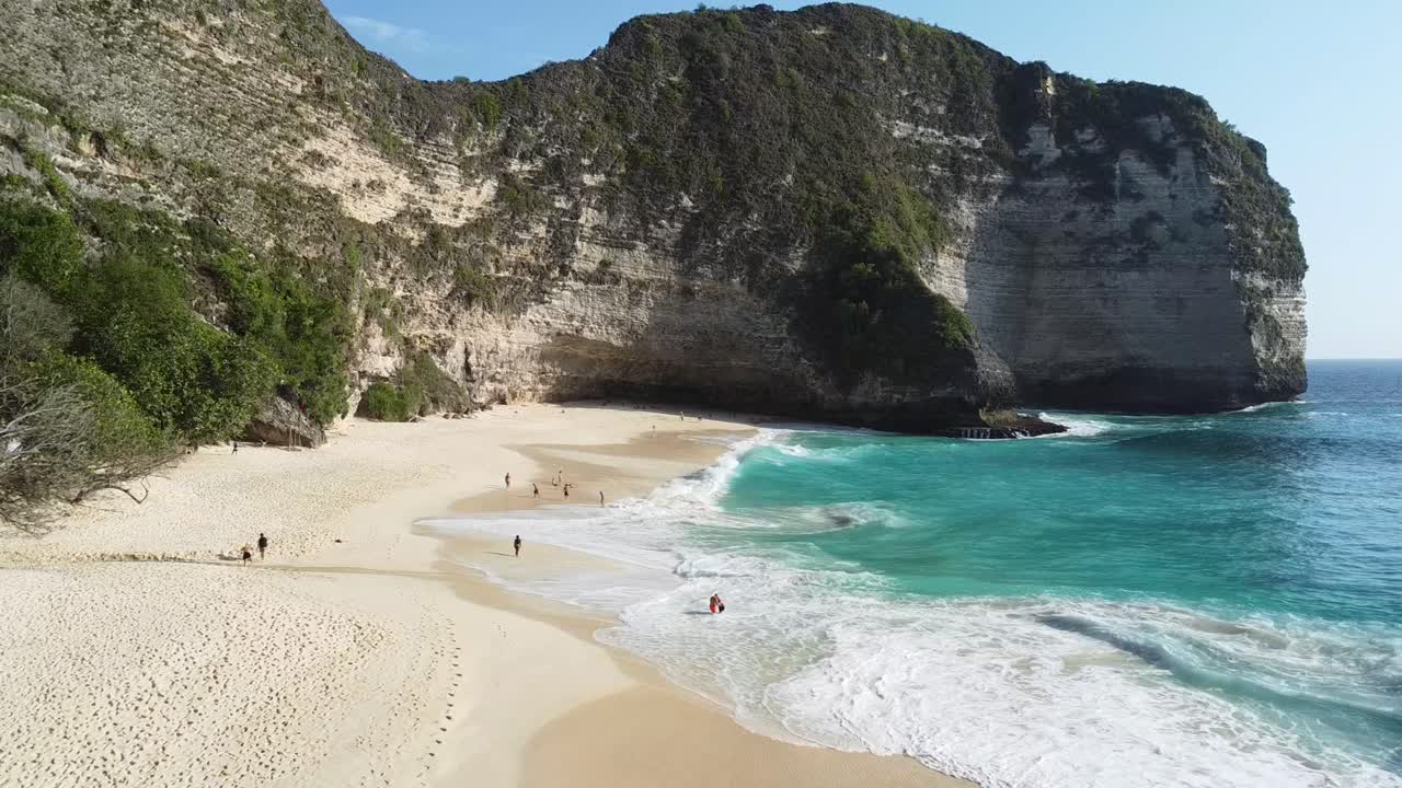 Drone panoramic dolly establishing view of Kelingking Beach, dynamic view of cliffs and clear water, Nusa Penida island with tourists enjoying sandy beach