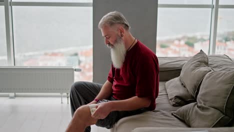 un hombre mayor con cabello gris y una barba espesa se venda la rodilla mientras está sentado en el sofá en un apartamento moderno con vistas al mar