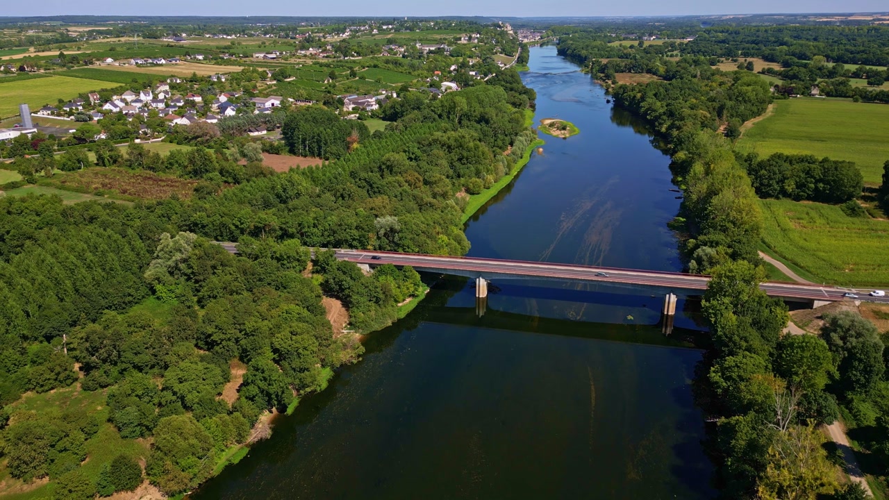 Aerial drone shot pulling back over the Vienne, showing a bridge with light traffic, a small island, and the town of Chinon in the background.